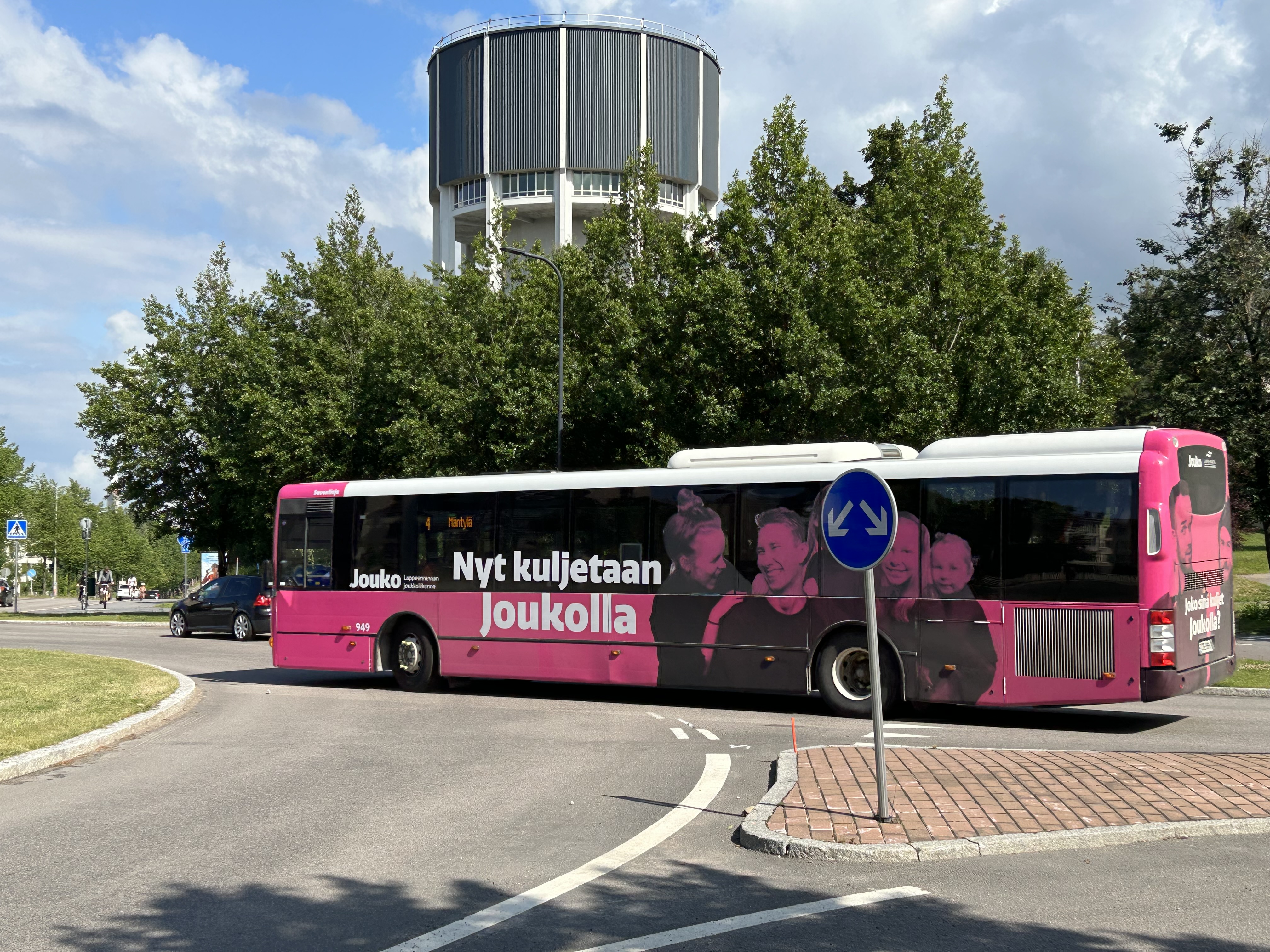 Pinkki Jouko-bussi liikenneympyrässä, vesitorni ja vihreitä puita taustalla. Bussissa teksti ”Nyt kuljetaan Joukolla”.